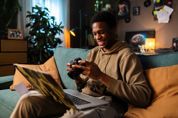 Carious delighted African American dark sinned black guy earing casual outfit sitting on sofa in cozy living room holding camera in hands observing photos while working online in laptop. - obrazy, fototapety, plakaty