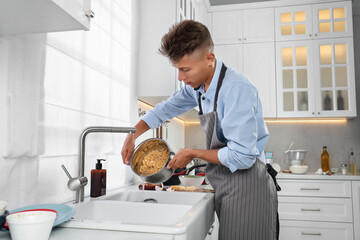 Man draining water from saucepan with pasta in messy kitchen. Many dirty dishware and utensils on countertop