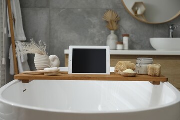 Wooden tray with tablet and spa products on bath tub in bathroom