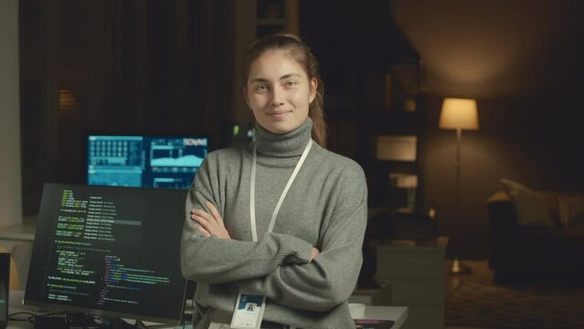 Medium slowmo portrait of young Caucasian female IT coordinator posing for camera with hands folded in dark programmers office at night
