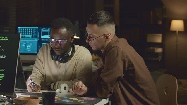 Waist up of two ethnically diverse male programmers working on new software at office desk late at night