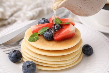 Pouring honey from jug onto delicious pancakes with strawberries, blueberries and mint at table, closeup