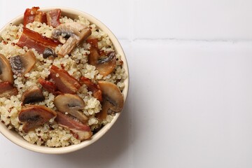 Tasty quinoa porridge with fried bacon and mushrooms in bowl on white tiled table, top view. Space for text