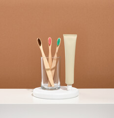 Several eco friendly toothbrushes with multicolored bristles stand in a glass on a white round stand. A tube of toothpaste stands on the table.