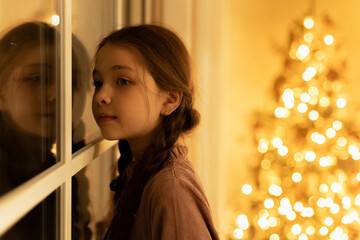 little girl with braids stand near window and look out, waiting santa claus with presents during christmas night
