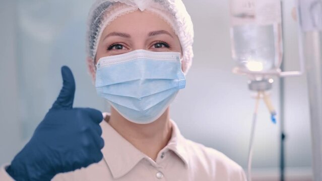 Cheerful nurse showing thumbs up. Woman in protective mask and surgical cap preparing IV bag for intravenous infusion in hospital. Close up of face.