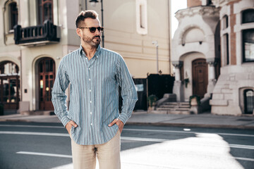 Handsome confident stylish hipster lambersexual model. Sexy modern man dressed in blue shirt and trousers. Fashion male posing on street background in Europe city at sunset. In sunglasses. Closeup - obrazy, fototapety, plakaty