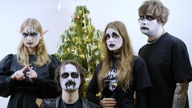 Four people stand in front of a decorated Christmas tree, each with dramatic black and white face paint, resembling gothic or fantasy characters. Two women wear black, with one having elf-like ears