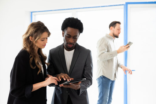 Blonde young beautiful woman talking with african americal real estate agent choosing house discussing house home future homeowners while handsome busy husband observing house potential clients.