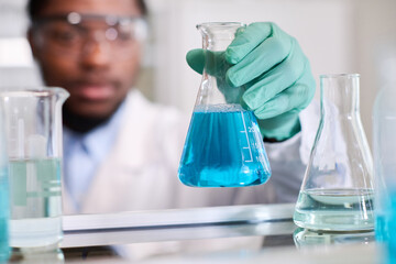 Close up shot of conical flask with blue liquid substance in hand of defocused African American...
