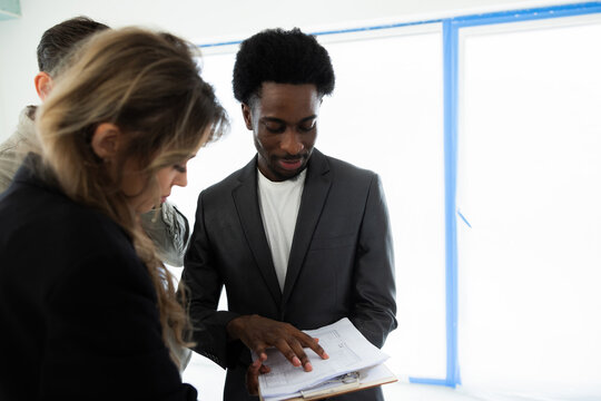 African american darl skinned real estate agent holding plan in hands showing to customers clients examning observing new plan of building standing at home house future investment. - Powered by Adobe