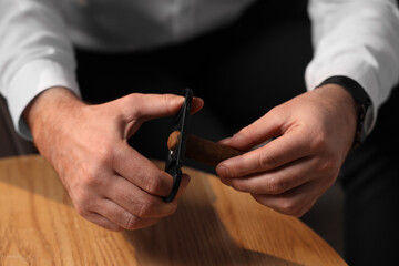 Man cutting tip of cigar at wooden table, closeup - obrazy, fototapety, plakaty