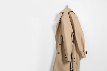 Beige trench coat hanging on hook rack on white wall, space for text