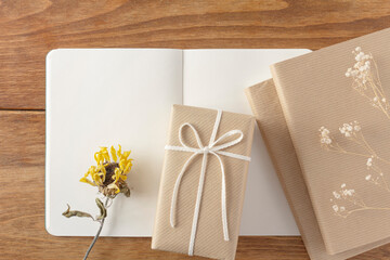 On a brown wooden table with an open notebook, a gift, a book, dried hazel flowers, and dried sunflowers.