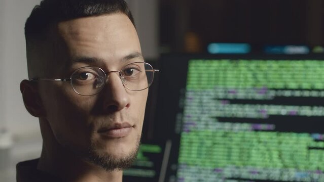 Medium closeup portrait of young Biracial male programmer in eyeglasses posing for camera while program coding on desktop computer in office at night