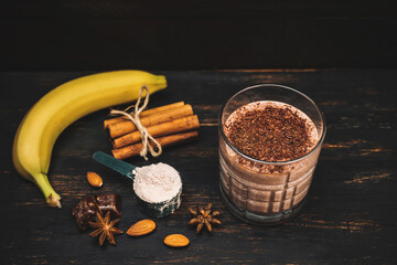 Milkshake smoothie, protein drink with grated chocolate in a glass on dark wooden board with bananas, protein powder in measuring spoon, star anise, almonds, cinnamon stick, chocolate pieces