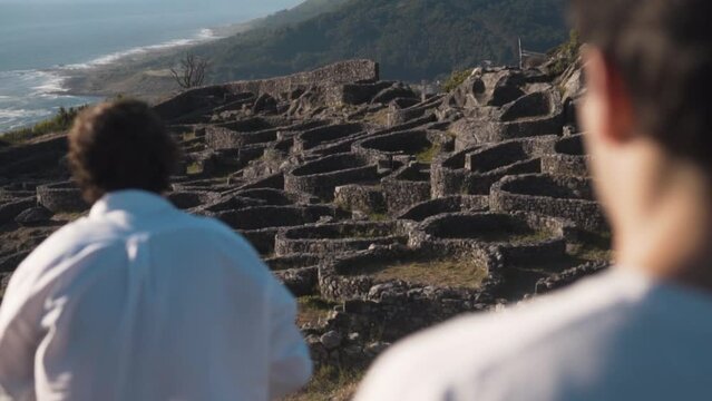 Sunlit summer eve on Santa Tecla, A Guarda. Two young men, one absorbed in the view, the other setting up a camera on a tripod. Ancient Castro, lush hills and Atlantic ocean provide a serene backdrop.