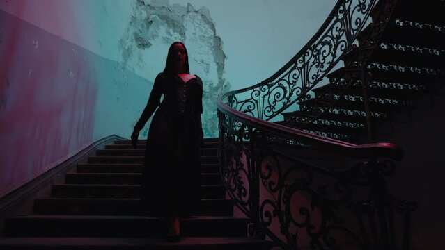A gothic woman in a black halloween outfit walks down the stairs. The hallway is lit by spooky red lamps, creating a horror movie vibe. It's nighttime indoors.