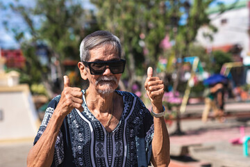 A cool hippie granddad making the thumbs up sign, gesturing with both his hands. Wearing shades and boho style shirt. Hanging out at the park. - obrazy, fototapety, plakaty