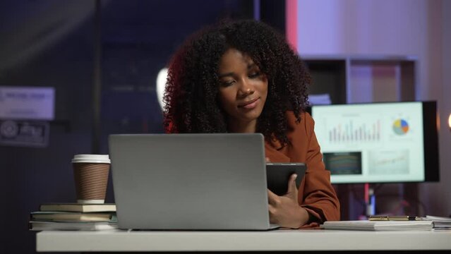 joyful African American office worker with an afro, talking on a mobile phone and writing on a notepad, with a stack of papers and a laptop on the desk.	
