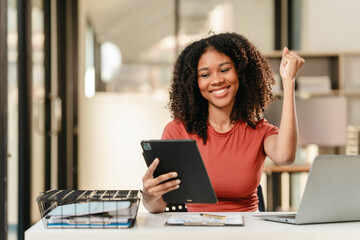 Portrait of joyful African American woman people with an afro hairstyle, working on a laptop at a desk with documents. - obrazy, fototapety, plakaty