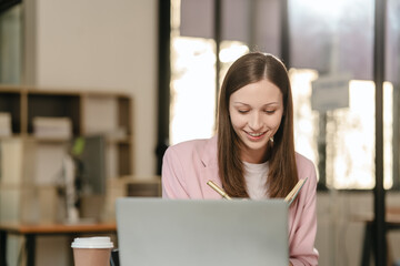 smiling Caucasian university student at a desk with a laptop, likely studying a subject in the...