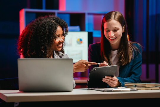 happy African American office worker with an afro and a Caucasian colleague collaborating over a laptop and documents at night.