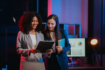 African American worker with an afro and a Caucasian worker discussing a clipboard in a vibrant office setting, possibly during overtime.