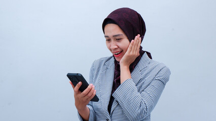 Asian woman in hijab formal outfit smiling and holding smartphone, isolated on gray background.