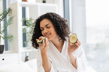 Close up portrait of young multinational female in robe holding two avocado halves in hands in...
