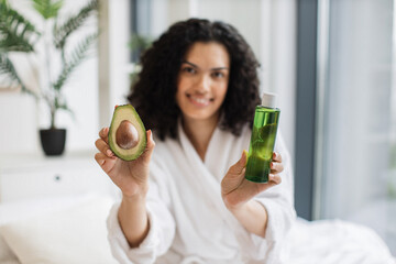 Focus on hands of African woman in after-shower outfit presenting avocado and cosmetic oil while sitting behind bed tray at home. Cheerful lady recommending natural ingredients in facial therapy.