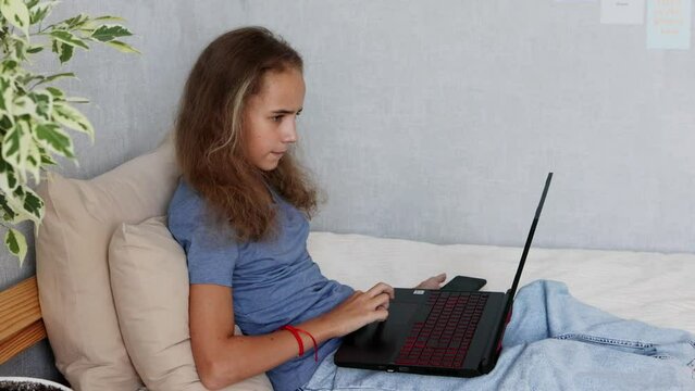 A cute blonde teenage girl is typing on a laptop while sitting on her bed in the bedroom