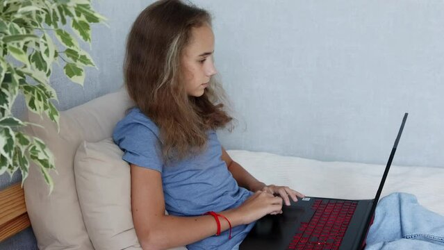 A blonde teenage girl is sitting on her bed typing on her laptop computer in her bedroom