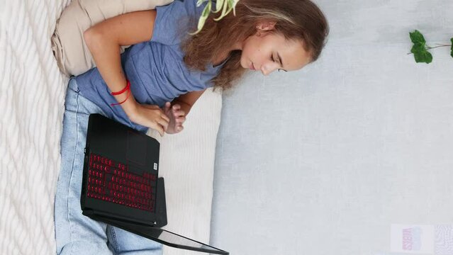 A pretty teenage girl with blonde hair is sitting on the bed in her bedroom and typing on her laptop