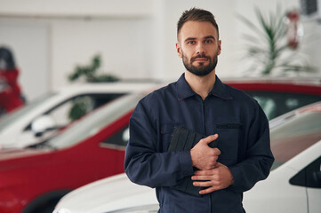 In blue colored uniform. Car repairman is in the garage with automobile