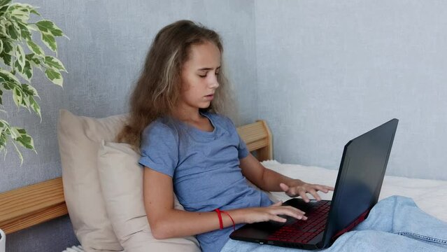 In her bedroom, a pretty blonde girl is sitting on the bed and typing on her laptop
