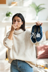 Disgusted beautiful woman holding stinky shoes in the living room at home.