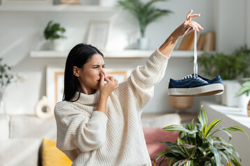 Disgusted beautiful woman holding stinky shoes in the living room at home.