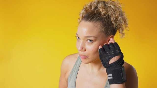 Studio shot of woman in boxing fitness clothing and sparring gloves streaming to wireless ear buds against yellow background
