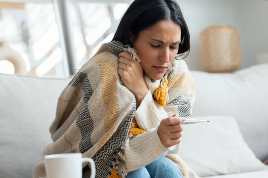 Tired woman with a cold taking her fever while lying with a blanket on the couch at home