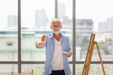 Cheerful senior man showing paintbrush and painting on canvas at home, Elderly man painting on a...