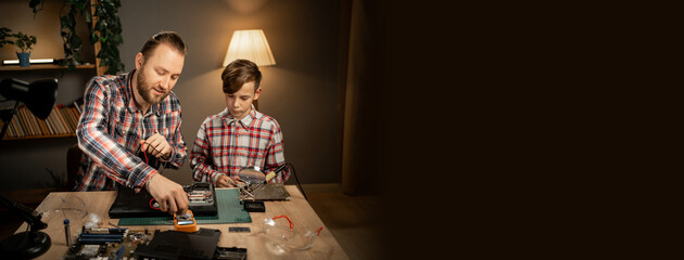 Father teaching his son to repair a broken laptop and fix the motherboard. Man and child soldering at home in the evening