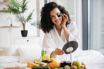 Portrait of young woman in after-shower outfit putting anti-aging gel patch under eye with help of cosmetic mirror placed on tray table in bedroom, looking at camera.