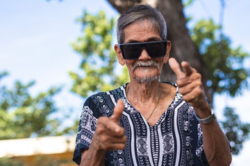 A cool hippie granddad pointing to the camera making finger gun gestures. Wearing shades and boho style shirt. Hanging out at the park. - obrazy, fototapety, plakaty