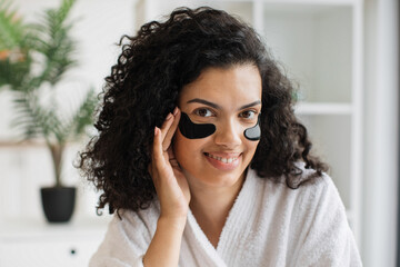 Close up view of lovely African female in white bathrobe applying pair of eye patches after...