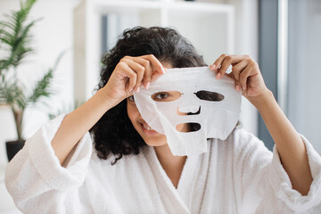Charming multinational female in white bathrobe looking through holes of cosmetic sheet mask cut...