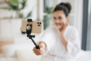 Focus on phone of relaxed adult person blogger in after-shower wear making media content while applying facial mask using makeup mirror from tray table while sitting on bed in studio room.