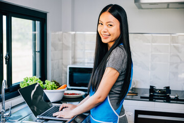 In the kitchen a smiling woman stands engrossed in cooking and using her laptop. Emphasizing the...