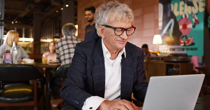 Adult Businessman Smiling While Working On His Laptop In Cafe. Behind Successful Man Visible Hardworking Waiter Taking Order From Customers. People Receiving Food. Working Day In Cafeteria.