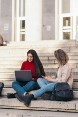 Two student girls sitting on the stairs of the college with a laptop and mobile.